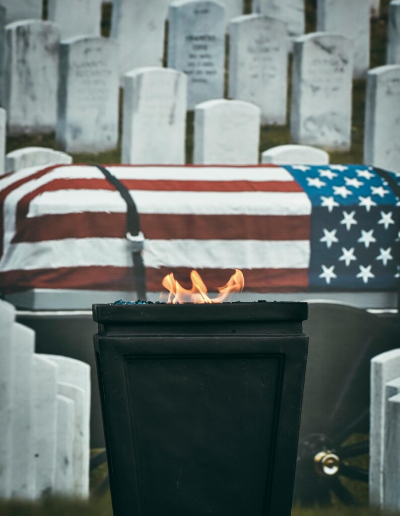 A solemn cemetery scene with an American flag draped coffin and eternal flame, honoring fallen veterans.